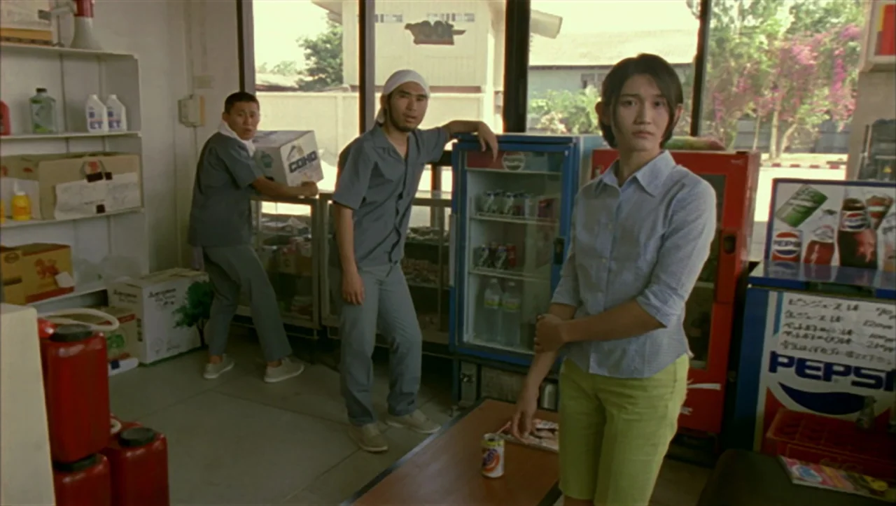 Wild Zero (1999) movie still: fullbody — A small retail shop features beverage coolers and stocked wooden shelves against bright windows. Three…