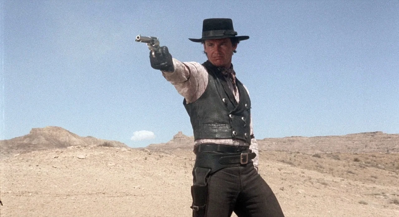 The Shooting (1966) movie still: medium — A dry desert landscape under a clear blue sky with rocky hills in distance. A man in western attire stands…