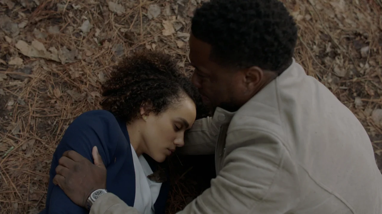 Die Hart (2023) movie still: medium — A dense forest floor is covered with dry pine needles and fallen leaves. A man crouches to support a woman…