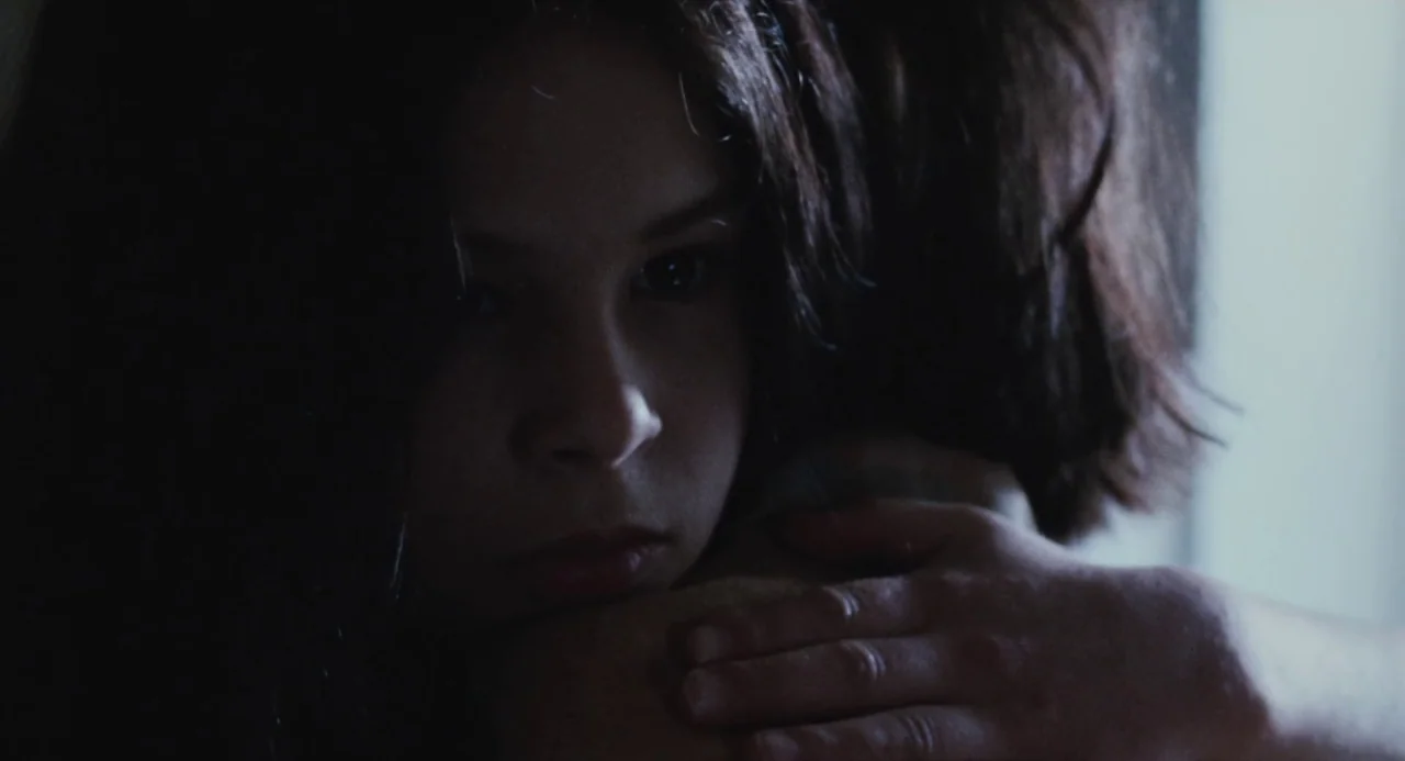 Martyrs (2008) movie still: closeup — A close-up view shows a girl resting her head on someone's shoulder indoors. Soft light hits the side of her…