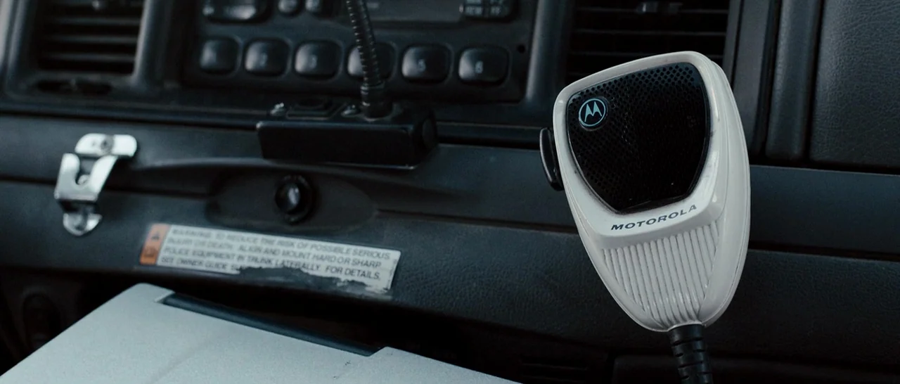 Cop Car (2015) movie still: closeup — The interior dashboard of a police cruiser features a mounted Motorola radio microphone. A warning label is…