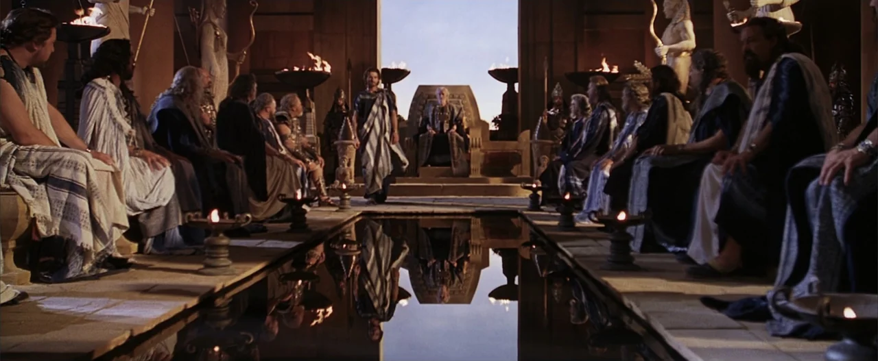 Troy (2004) movie still: wide — A stone hall features a central reflecting pool flanked by rows of seated figures. A lone man walks between the…