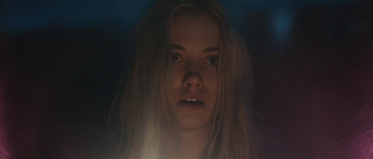 Starfish (2018) movie still: closeup — A woman stands in extreme darkness with only her face visible in the light. She looks directly forward into…