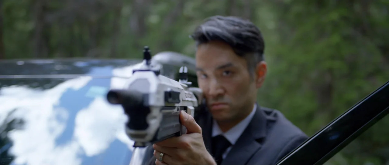 La presencia (2012) movie still: medium — An Asian man in a suit aims a firearm in a forested area. A dark vehicle sits in the background behind him.