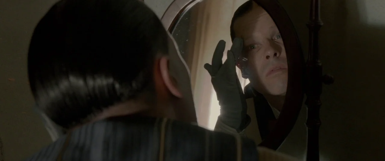Lawless (2012) movie still: closeup — A man looks into a small oval mirror mounted on a dark wooden frame. He is wearing a grey glove and adjusts…