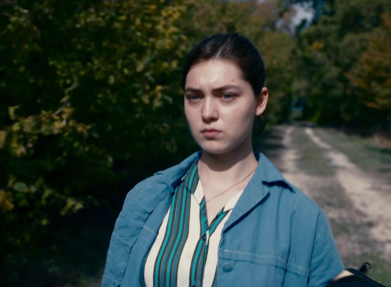 Happening (2021) movie still: medium — A woman stands on a dirt path flanked by dense green foliage and trees. The subject looks forward against a…