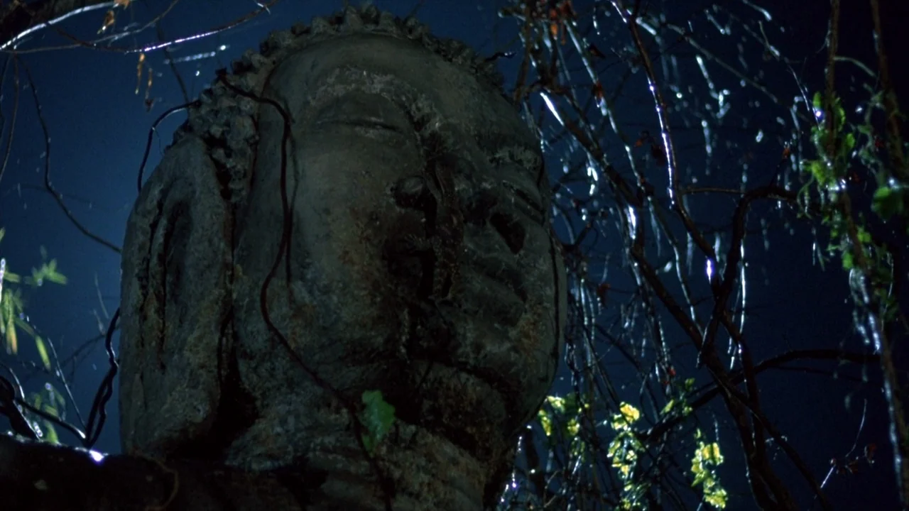 Platoon (1986) movie still: closeup — A weathered stone Buddha head sits outdoors at night, partially obscured by dark, wet tree branches. Rain…