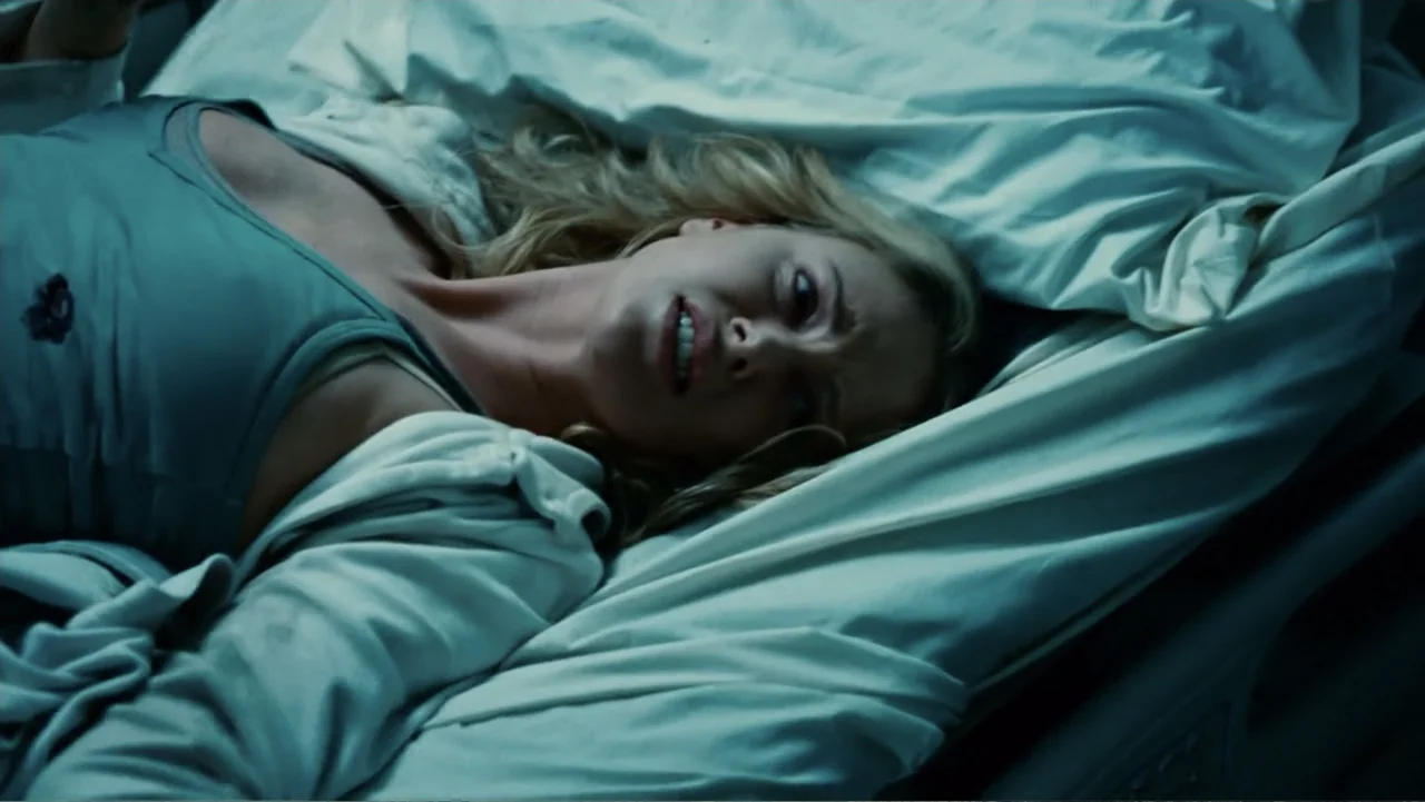 Hancock (2008) movie still: medium — A hospital room with white linens and clinical features is dimly lit. A blonde woman in a tank top lies on the…
