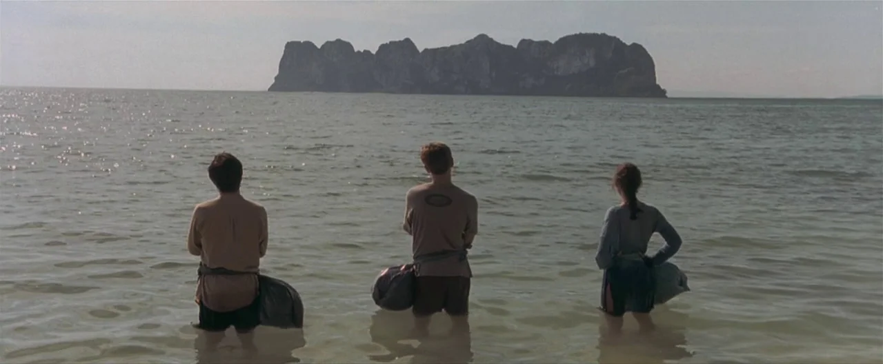 The Beach (2000) movie still: wide — A calm, expansive body of water separates the shoreline from a large, jagged limestone island. Three people…