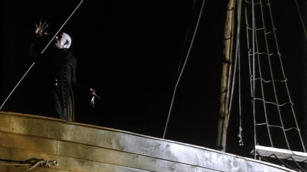 Shadow of the Vampire (2000) movie still: wide — A dark wooden ship deck at night with a tall mast and rigging. A pale figure in a black coat stands…