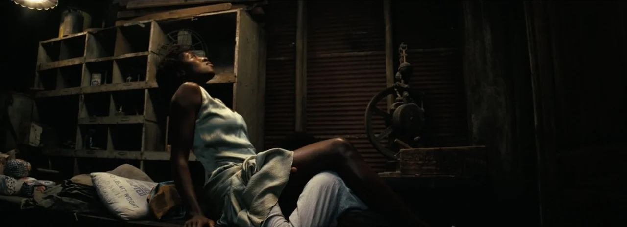Sinners (2025) movie still: medium — A dark wooden shack interior contains a single bed and an industrial flywheel mechanism. A young woman in a…