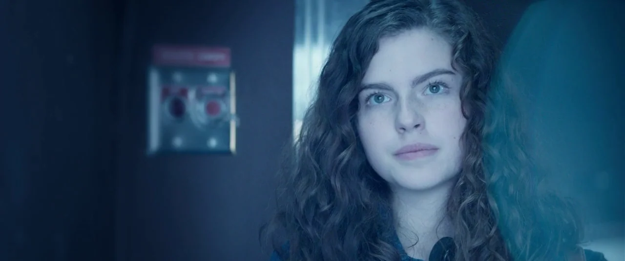 Steve Jobs (2015) movie still: closeup — An interior space features a dark wall with a mounted metallic alarm switch plate. A young woman with long…