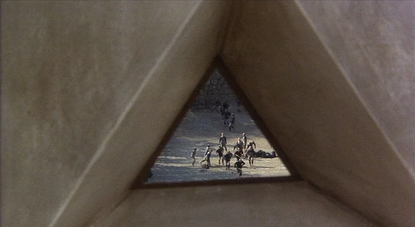Kagemusha (1980) movie still: wide — A view through a triangular stone opening reveals an outdoor courtyard with a dirt surface. A crowd of people…
