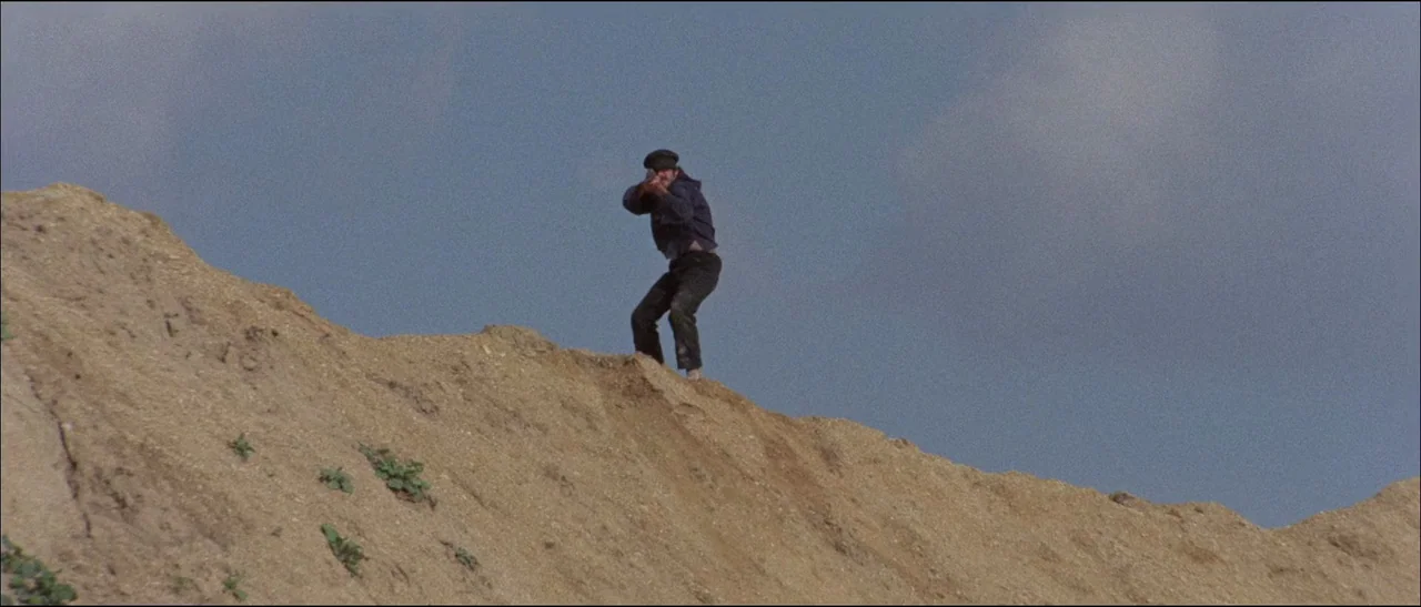 The Grand Duel (1972) movie still: wide — A man stands atop a steep sandy ridge against an overcast pale sky. The man is holding a rifle and…