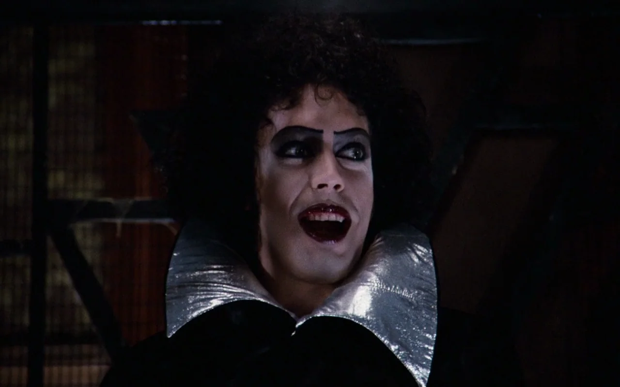 The Rocky Horror Picture Show (1975) movie still: closeup — A dark indoor space features a subject with dramatic makeup and a silver collar. The…