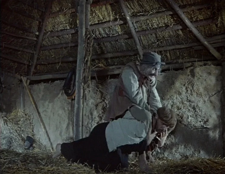 Viy (1967) movie still: fullbody — A rustic barn with a thatched roof and rough clay-plastered wall interior. An older man grips a younger man from…