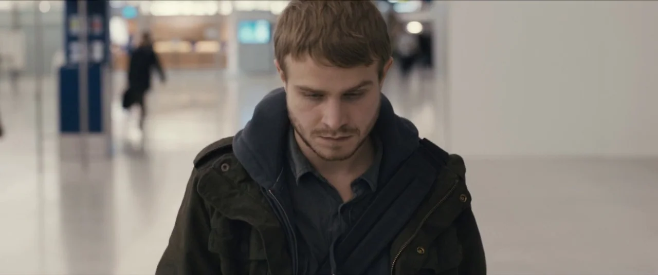 Simon Killer movie still: medium — A sterile, brightly lit airport terminal with blurred signage and reflective flooring. A young man walks through…
