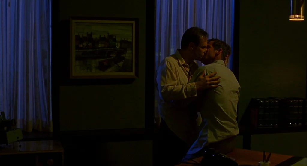 Far From Heaven (2002) movie still: medium — A dimly lit office at night features two large windows covered by sheer white curtains. Two men embrace…