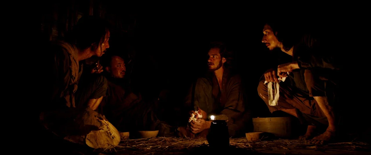 Silence (2016) movie still: wide — A dark, cramped interior with straw scattered across the wooden floor. Four men sit huddled around a small…