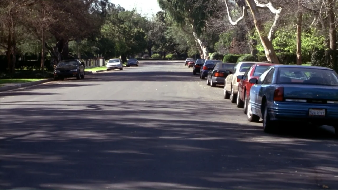 Music from Another Room (1998) movie still: establishing — A tree-lined suburban street with parked cars stretching into the distance under clear…