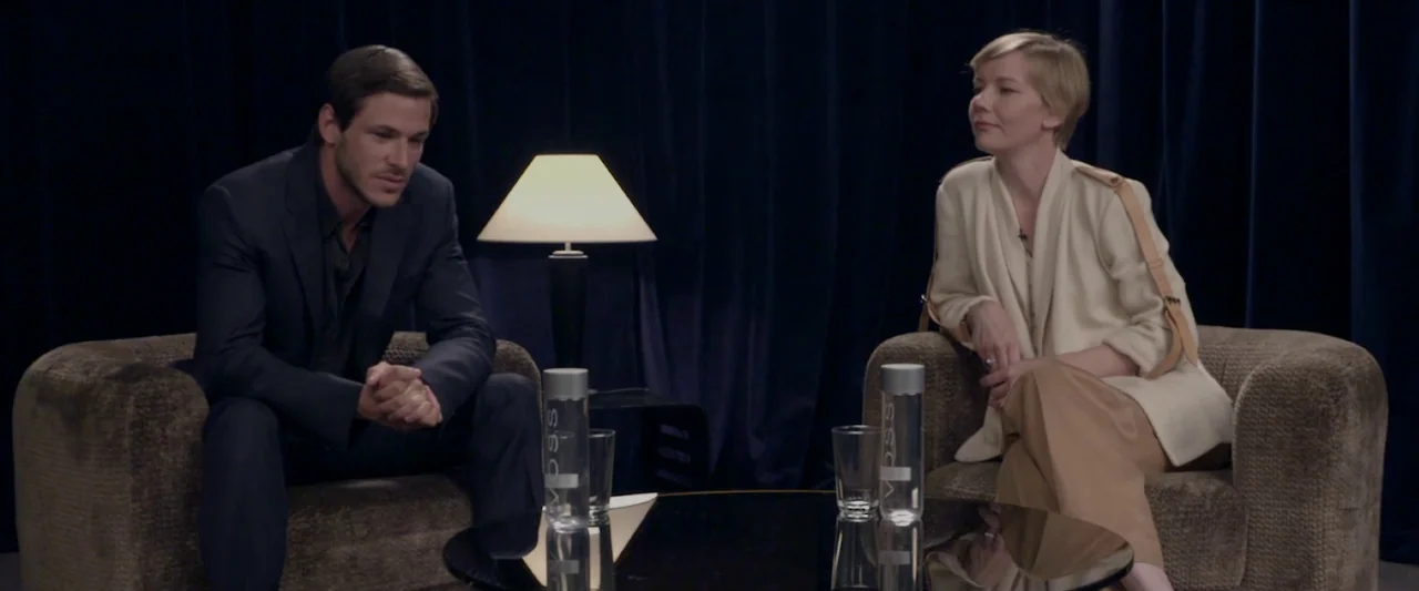 Sibyl (2019) movie still: medium — A dark interview set features two individuals sitting in armchairs against blue curtains. A small table with a…