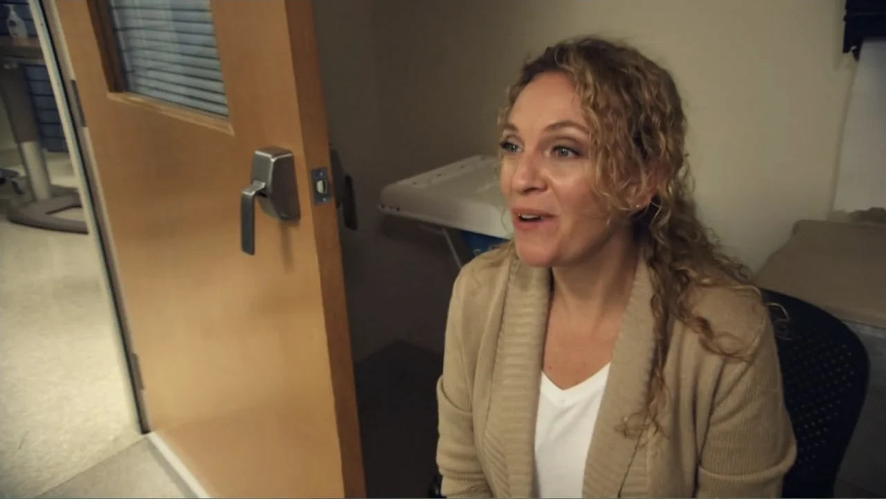 Suited (2016) movie still: medium — A medical exam room features plain walls and a wooden door left slightly ajar. A woman with curly blonde hair…