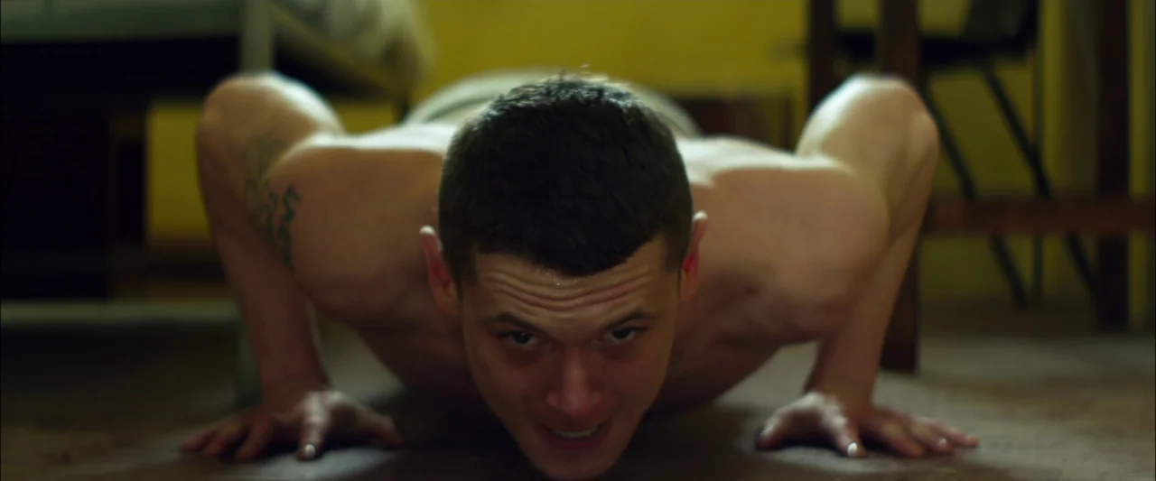 Starred Up (2013) movie still: medium — A small, stark interior room with mustard-yellow walls and dark floorboards. A shirtless young man performs…