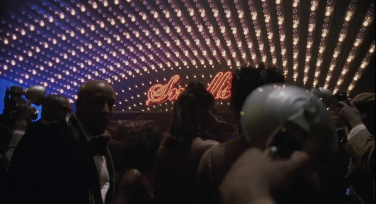 American Gangster (2007) movie still: medium — A crowded indoor space features a ceiling lined with many glowing incandescent light bulbs. People…