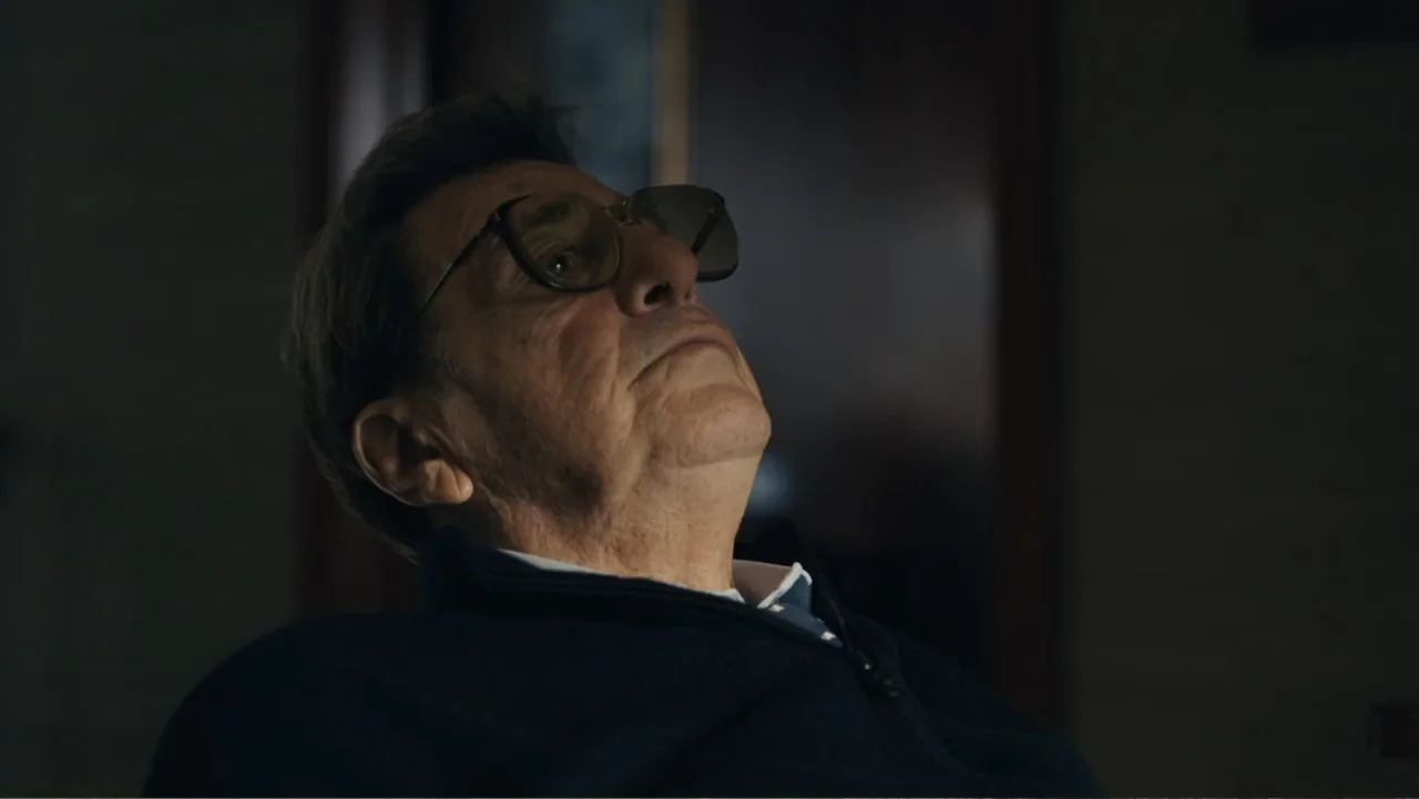 Paterno (2018) movie still: closeup — A dimly lit indoor space with blurred background architectural details. An older man with glasses tilts his…