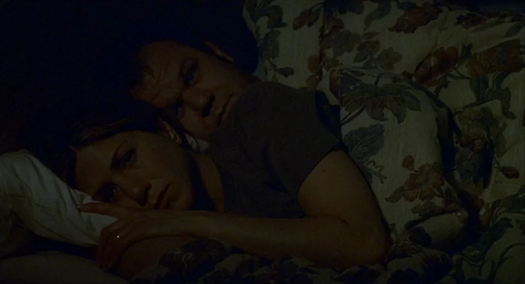 The Good Girl (2002) movie still: medium — A dimly lit room features a bed with a floral patterned duvet. A man and a woman lie together in the dark…