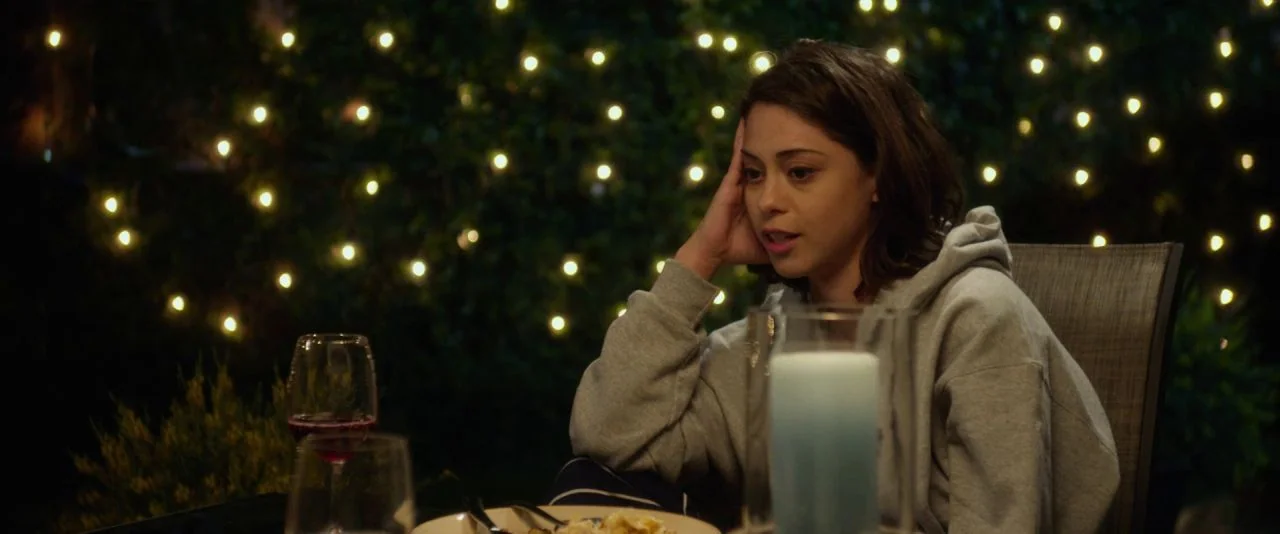 Night Owls (2015) movie still: medium — An outdoor evening setting features a table with a wine glass and candle. A young woman in a hoodie sits at…