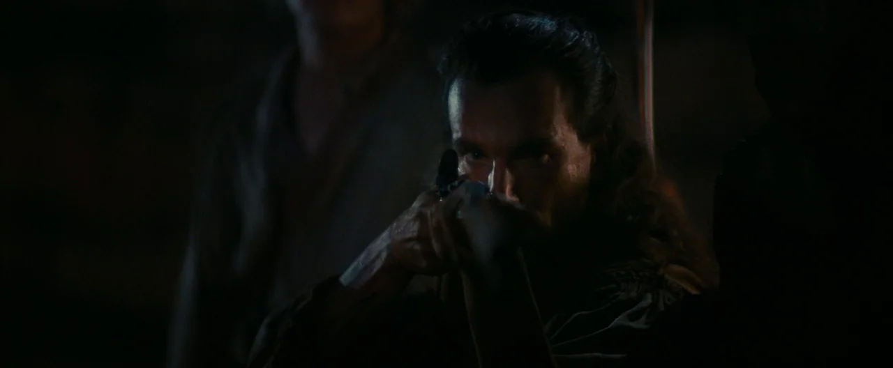 The Last of the Mohicans (1992) movie still: closeup — A dark, dimly lit environment with indistinct shadows surrounding a central male figure. The…