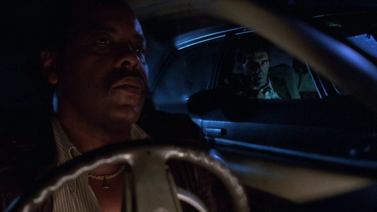 Repo Man (1984) movie still: medium — Two men sit inside a dimly lit car at night during a rainstorm. The driver focuses ahead while the passenger…