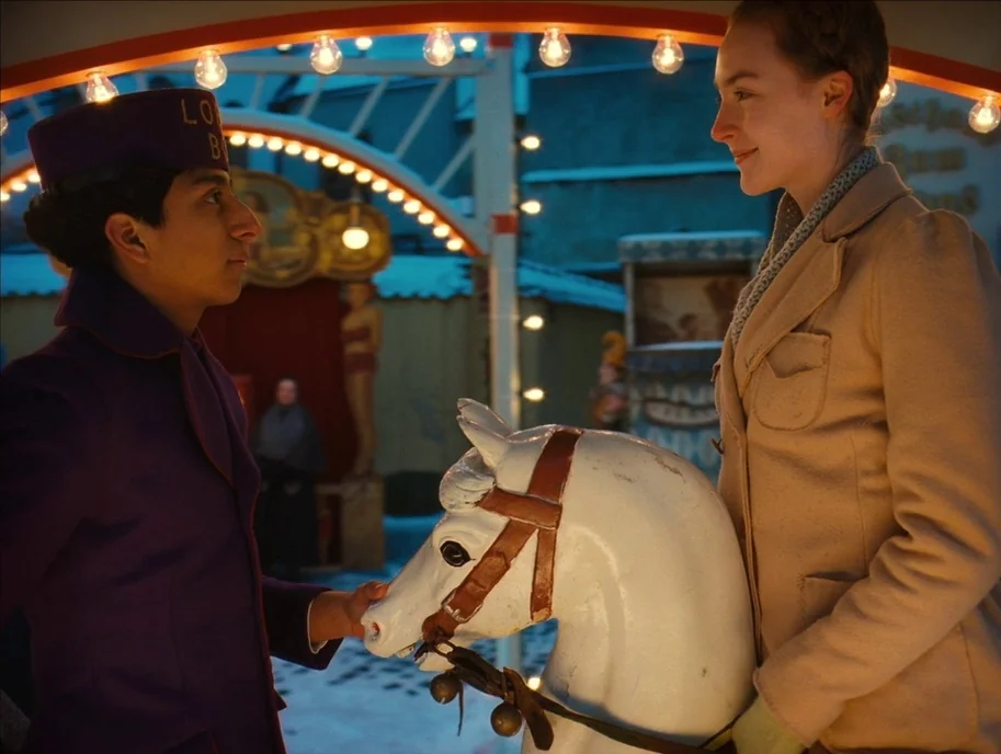 The Grand Budapest Hotel (2014) movie still: medium — A snowy outdoor carnival ground features a carousel at twilight with strings of lights. A…
