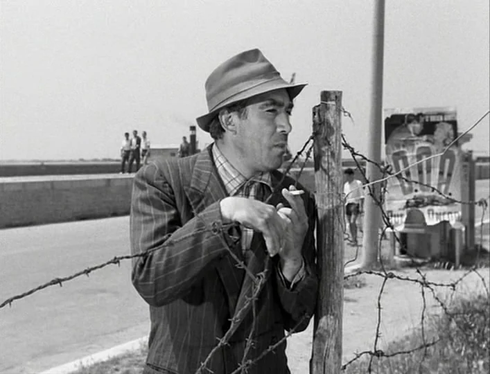 La Strada (1954) movie still: medium — A man stands outdoors next to a barbed wire fence near a paved road. A poster is attached to a post in the…