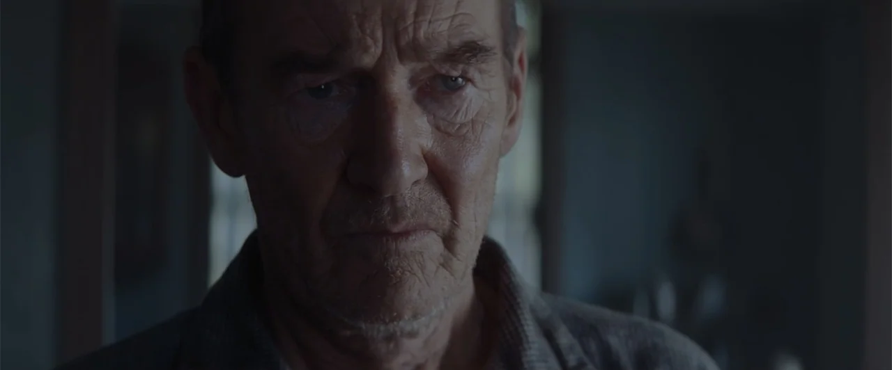 My Neighbor Adolf (2022) movie still: closeup — An interior space with a window providing soft, diffuse backlighting. An elderly man stares directly…