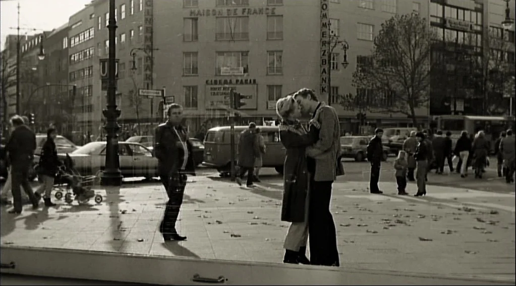 Wings Of Desire (1987) movie still: fullbody — A bustling Berlin city street scene with pedestrians, cars, and a subway entrance visible. A couple…