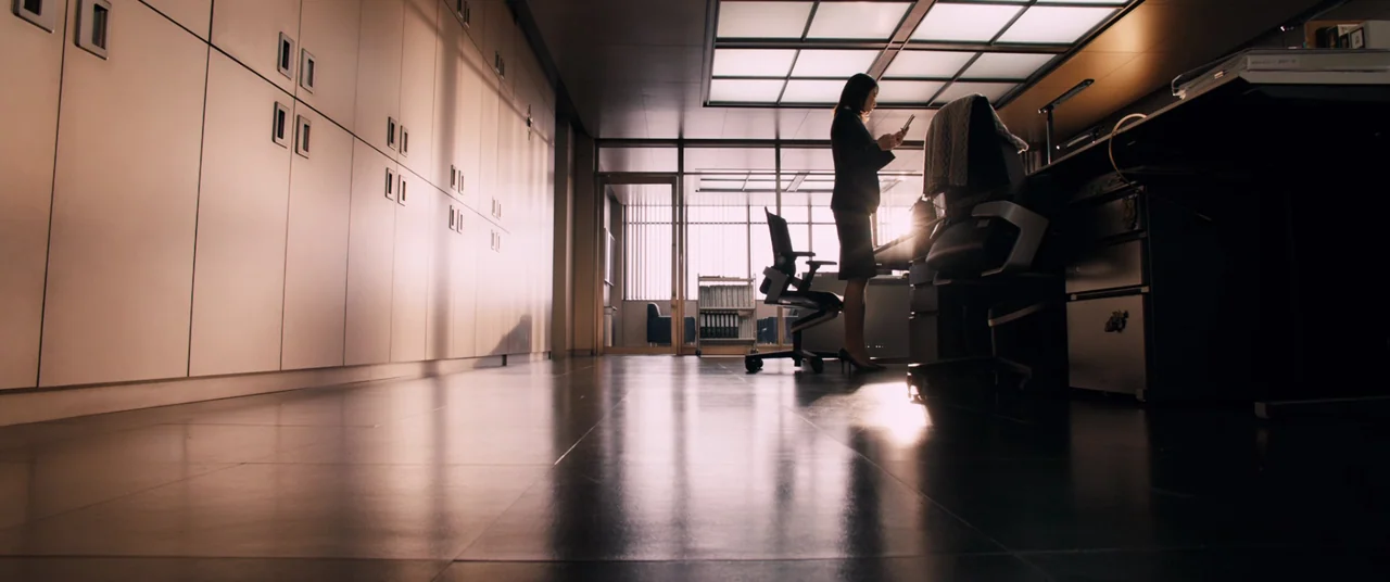 Shin Ultraman (2022) movie still: wide — A modern workspace features rows of metal cabinets and a large desk surface. A woman stands in silhouette,…