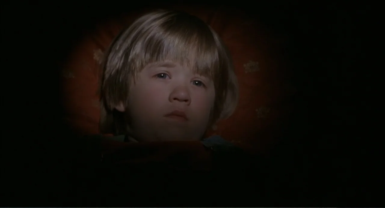 Bogus (1996) movie still: closeup — A dark bedroom interior features a small child illuminated by a dim spotlight. The boy lies in bed while facing…