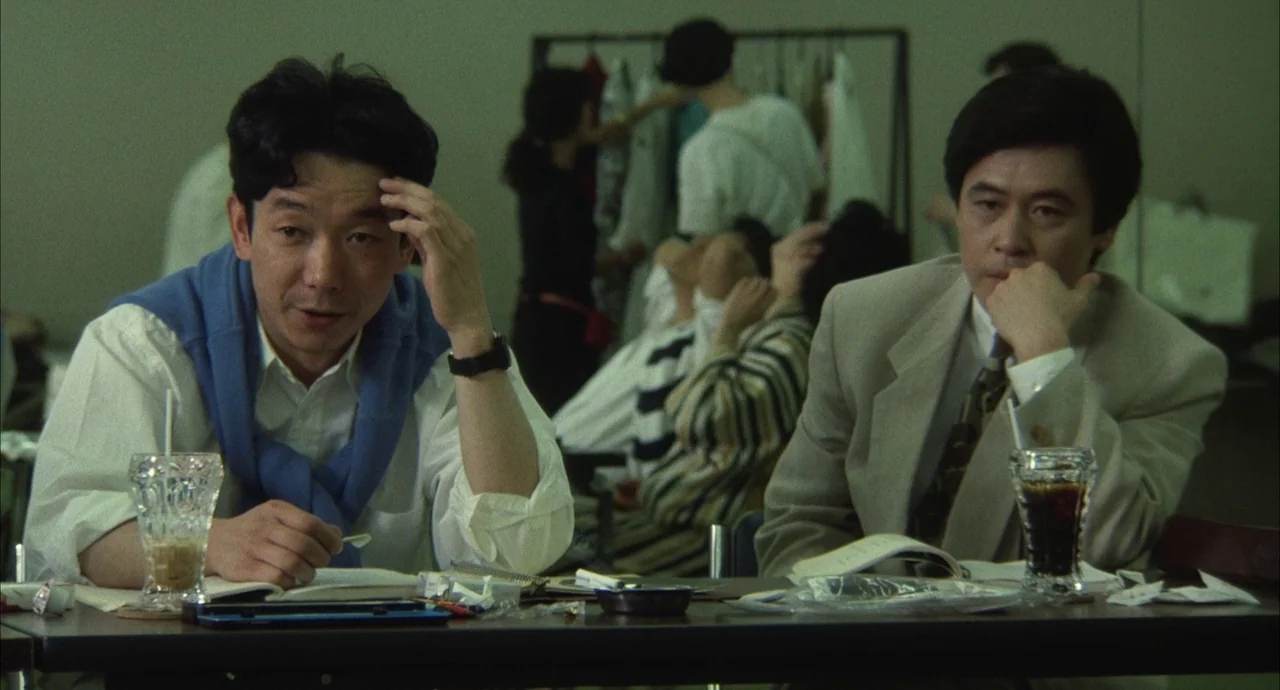 The Discarnates (1988) movie still: medium — An indoor space features a table with drinks and papers in the foreground. People congregate near a…
