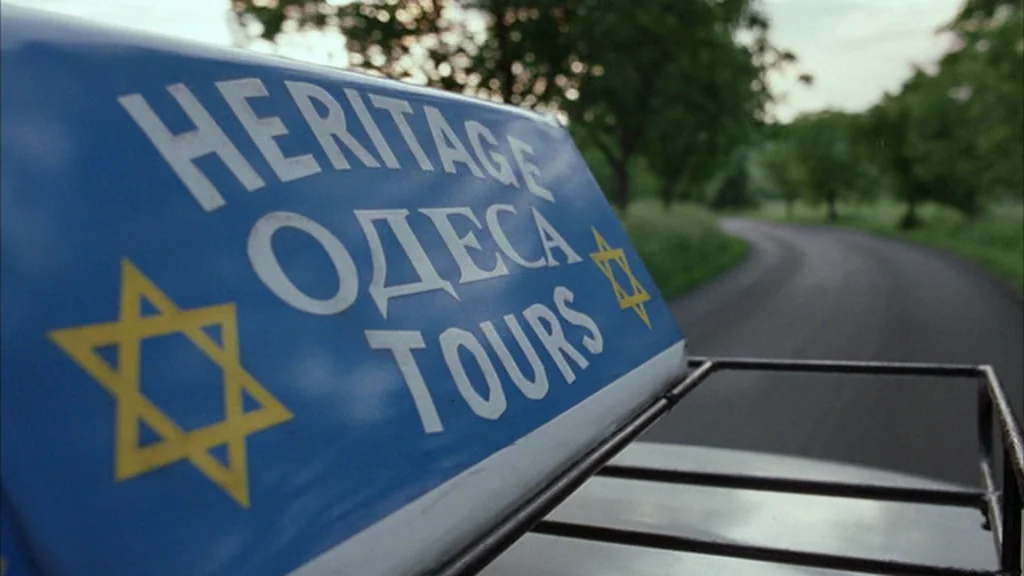 Everything Is Illuminated (2005) movie still: closeup — A blue sign reading Heritage Tours sits on the roof of a vehicle. The car travels along a…