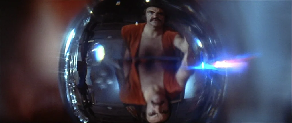Zardoz (1974) movie still: closeup — A reflective spherical object captures the distorted image of a man. The subject stares into the surface while…