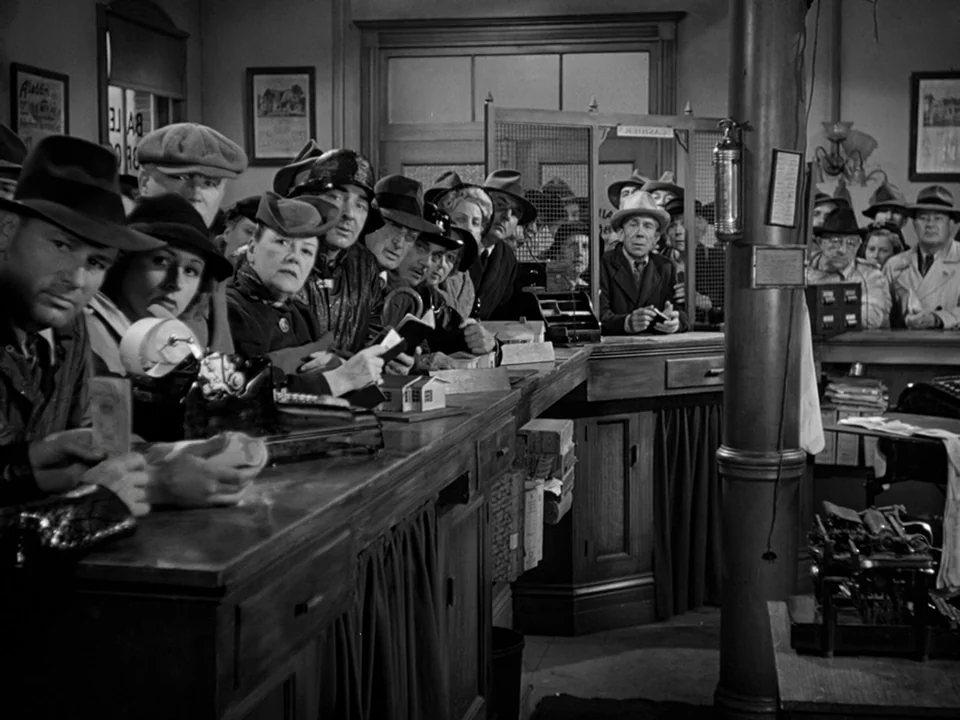 It’s A Wonderful Life (1946) movie still: medium — A crowded bank lobby features a long wooden teller counter with barred windows. A line of people…
