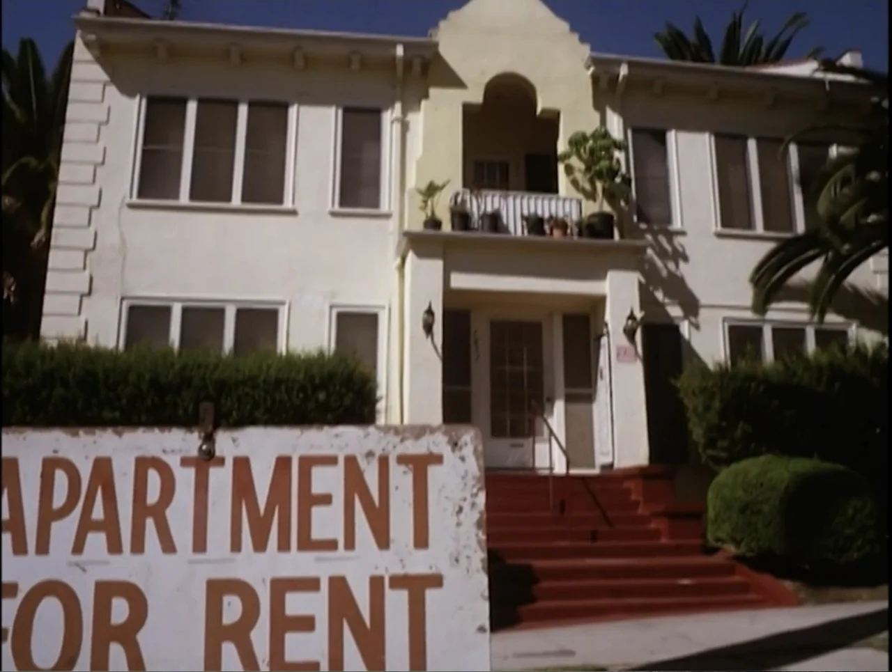 Apartment 12 (2001) movie still: wide — A light-colored stucco apartment building features red entrance steps and a second-floor balcony. In the…