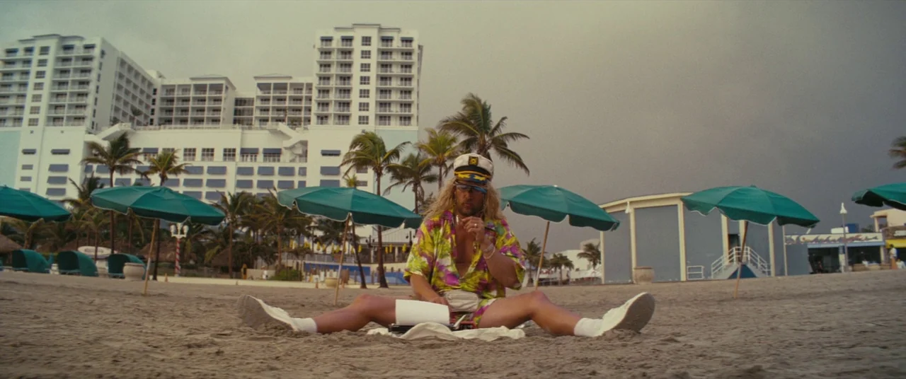 The Beach Bum (2019) movie still: fullbody — A sandy beach features multiple green umbrellas and a large hotel complex behind. A man sits on the…