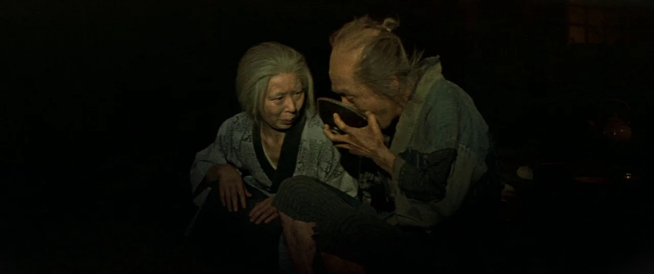 Ballad Of Narayama (1958) movie still: medium — Two elderly people sit together in a dark, dimly lit, sparse traditional structure. The man drinks…