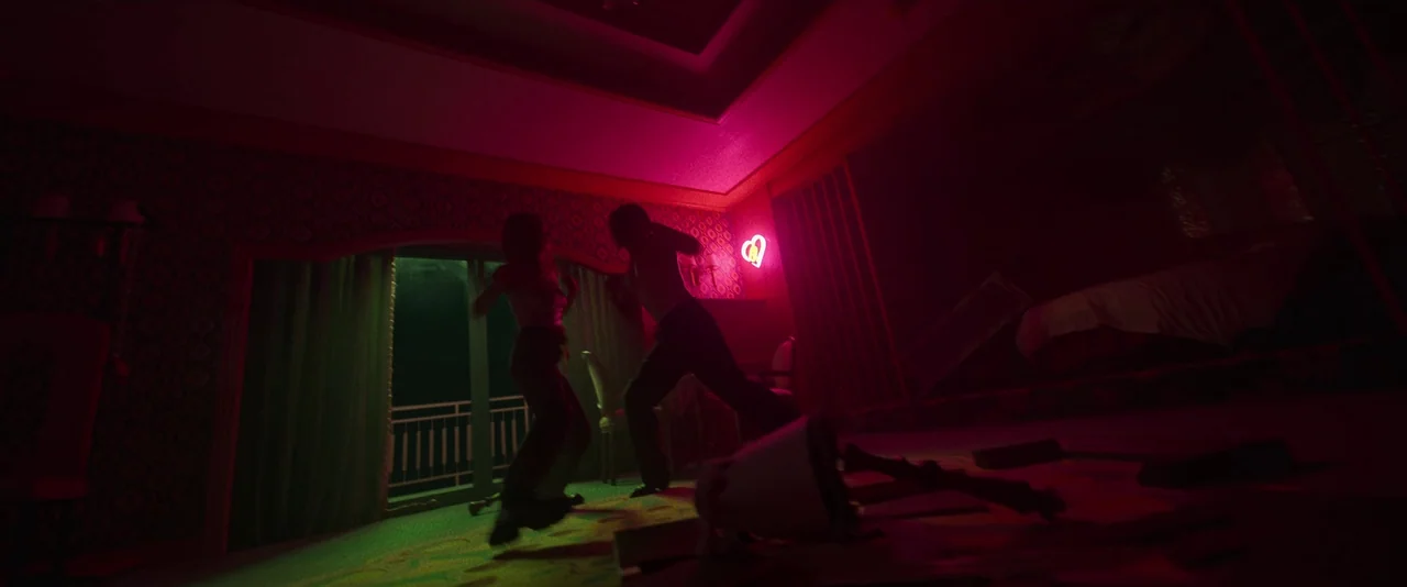 Ballerina (2023) movie still: wide — A dimly lit space features patterned wallpaper and a glowing neon heart sign on the wall. Two people stand in…