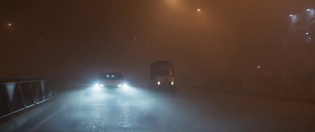 The Wedding Guest (2018) movie still: wide — A dark bridge is covered in a thick, murky orange fog at night. A car and a small three-wheeled vehicle…