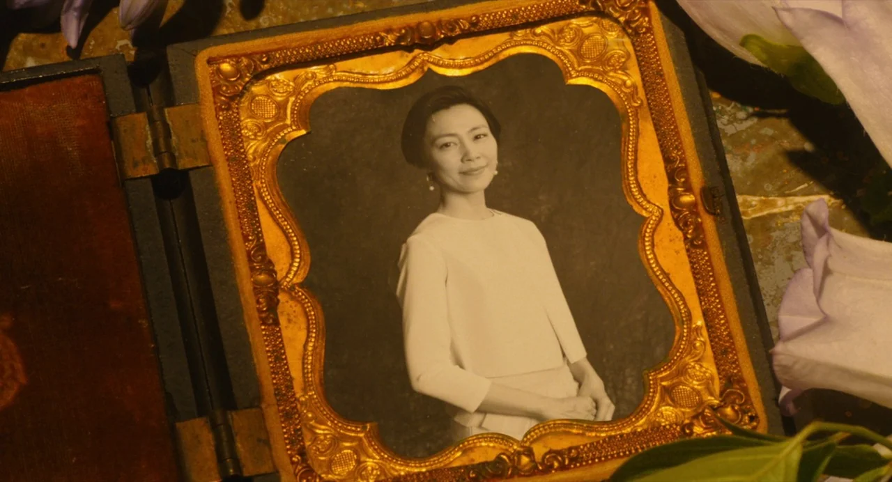 Diner (2019) movie still: closeup — An antique photograph in an ornate gold frame sits on a table. The portrait depicts an East Asian woman with…