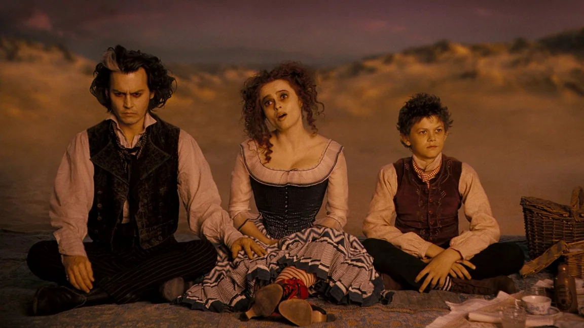 Sweeney Todd: The Demon Barber of Fleet Street (2007) movie still: medium — Three individuals sit in a line on a fabric blanket upon a sandy beach.…
